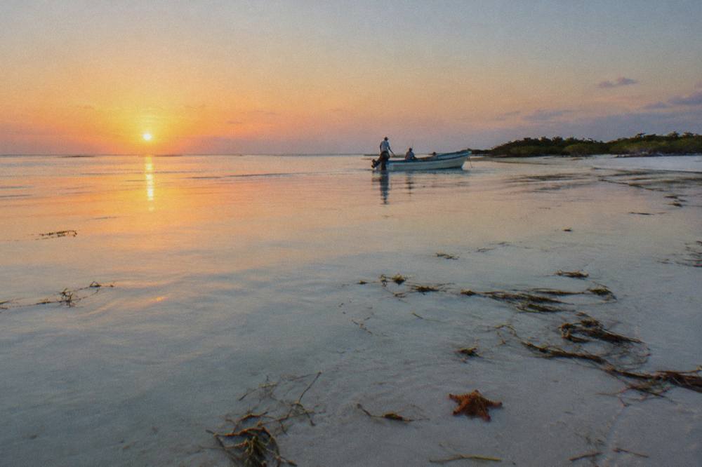 A small open boat crossing calm turquoise water toward a distant sandbar at sunrise