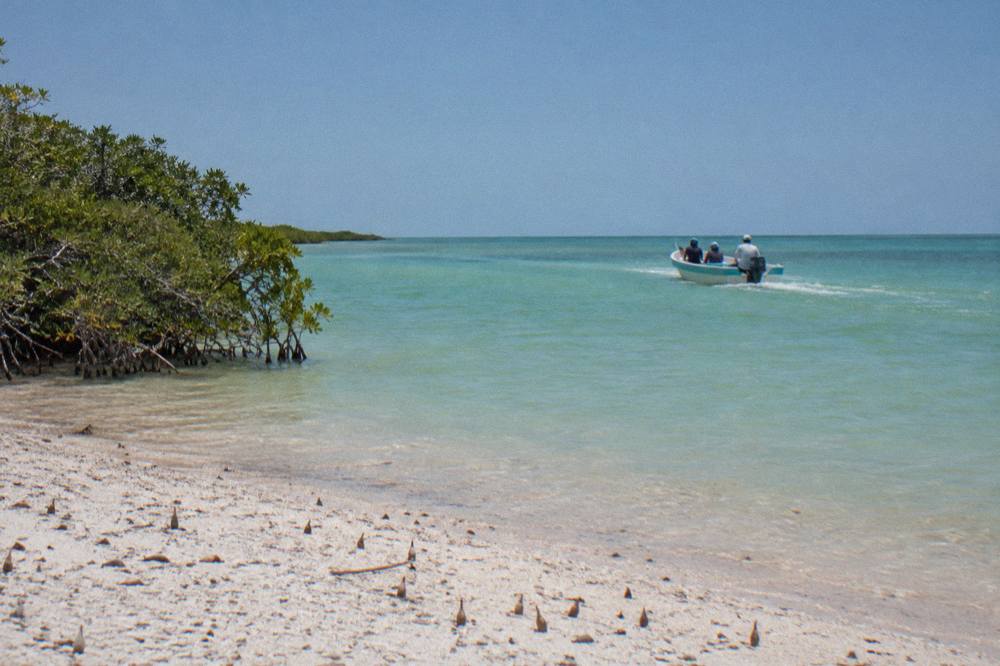 A small open boat crossing calm green-blue shallows toward open water at midday