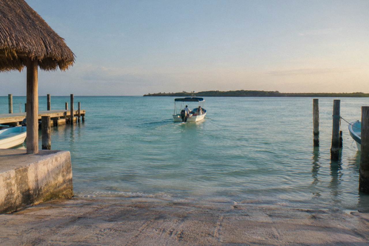 A small open boat crossing calm turquoise water near a low green island coastline under a bright sky