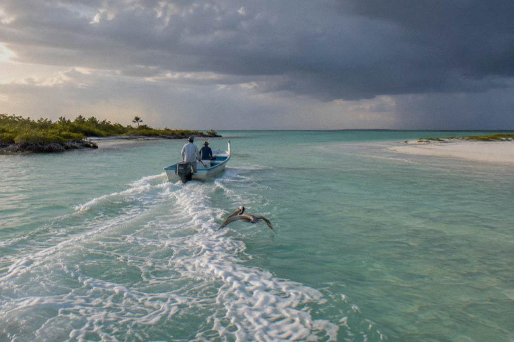 A small boat cruising turquoise shallow water past a sandbar toward Holbox