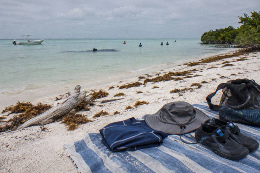 Rash guard, hat and water shoes folded on a beach towel beside a mesh packing bag